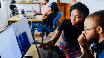 A woman overlooking a man using a computer.