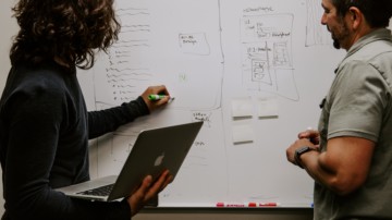 two people writing on a whiteboard, one is holding a laptop