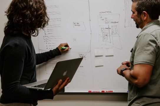 two people writing on a whiteboard, one is holding a laptop